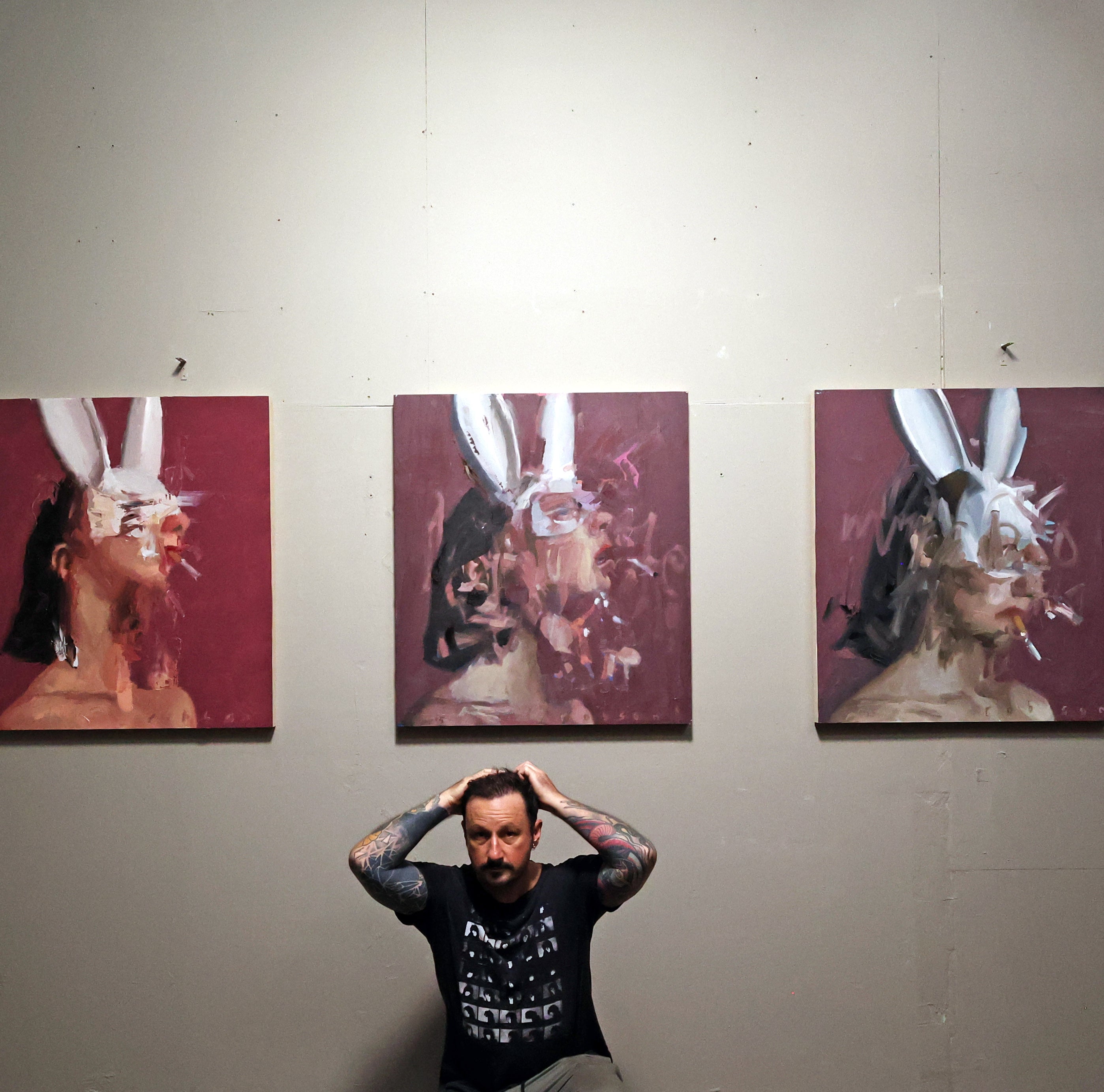 Artist seated beneath a three-panel portrait triptych on a gallery wall